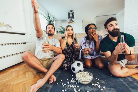 Very excited friends having fun by watching football match, drinking beer and eating popcorn at homeの写真素材