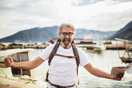 Smiling tourist mature man standing with map and backpack near the sea.の写真素材