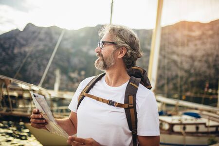 Smiling tourist mature man standing with map and backpack near the sea.の写真素材