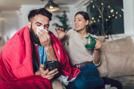 Cheerful romantic couple is sitting on sofa under cozy plaid and smiling. They are drinking tea.の写真素材