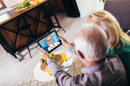 Senior couple at home holding digital tablet during video call with family doctorの写真素材