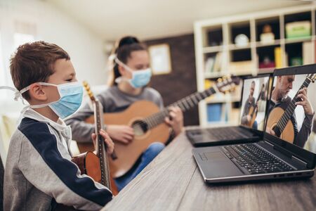 Boy and girl, wear protective masks, playing acoustic guitar and watching online course on laptop...