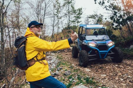 Middle age traveler in raincoat and backpack hitchhiking four-wheelers in forest.