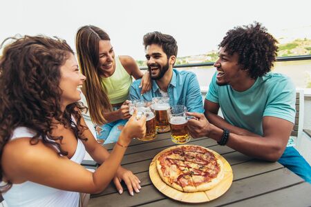 Friends enjoying pizza. Group of young cheerful people eating pizza and drinking beer while sitting at the bean bags on the roof of the buildingの写真素材