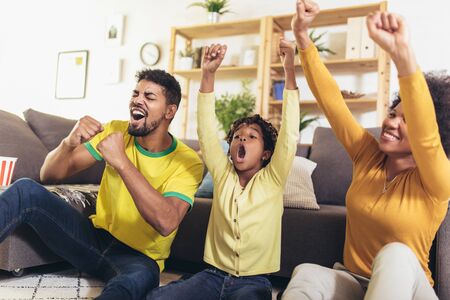 Happy African American family of three watching tv and cheering sport games on sofa at homeの写真素材