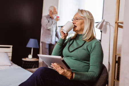Senior couple in bedroom. Woman using a tablet, man drinking wine.の写真素材