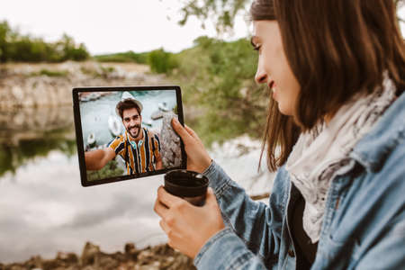 Young woman on camping near the river drinking tea an make video call on digital tablet.