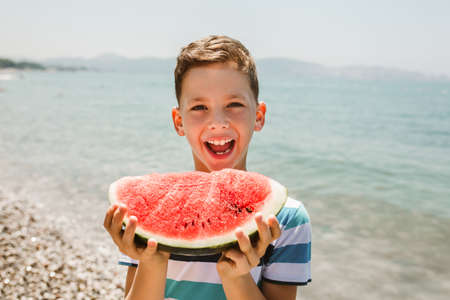 Cheerful child eating juicy watermelon. Kids emotions boy eating watermelon on the background of the sea, the beach, the sea coast.の写真素材