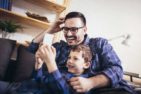 Young father embraces little son family sitting on couch at home using smart phone feels happy cheerful laughing on funny videos.の写真素材