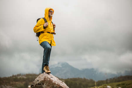 Middle age man traveler in raincoat and backpack enjoying view of mountains.の写真素材