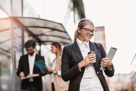 Young happy businesswoman holding digital tablet and coffee to go outside of modern building, businesspeople in background.の写真素材