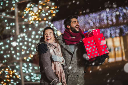 Young couple in the city centre with holiday's brights in background. Couple holding gift in a Christmas night.の写真素材