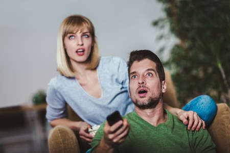 Young couple sitting together on a sofa at home watching television, joyfully smiling enjoying a...