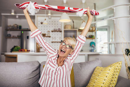Elderly woman emotionally watching soccer on tv and celebrating victory at home.の写真素材