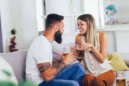 Young happy couple communicating while sitting on the sofa and drinking beerの写真素材