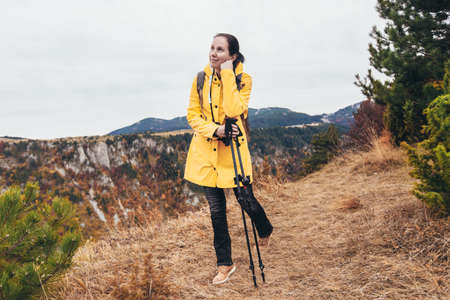 Happy mid age woman with backpack and raincoat standing on top of the mountain