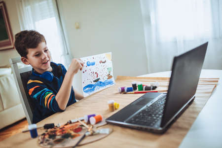 Focused boy painting and watching online course on laptop while practicing at home. Online training,...