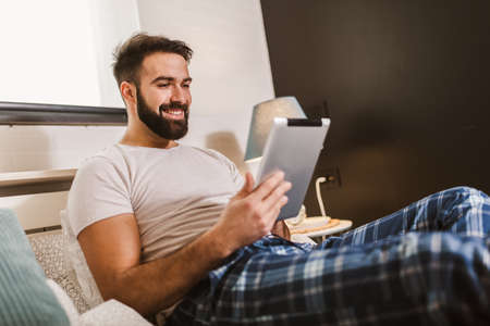 Handsome young man in pajamas using a digital tablet while lying in bed in the morning