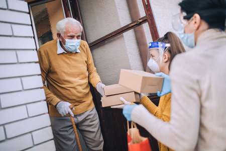 Young female volunteers in mask gives an elderly man boxes with food near his house. Quarantined, isolated. Coronavirus covid-19. Donationの写真素材
