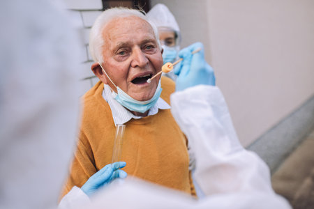 Doctors in a protective suit taking swab from a senior man to test for possible coronavirus infection. Taking corona virus test sample at home concept, senior man in quarantine.の写真素材