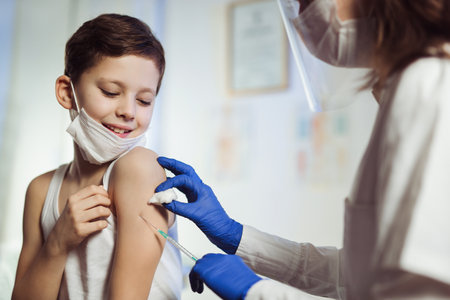 Pediatrician makes vaccination to cute Caucasian boy.They are wearing a protective face masks.