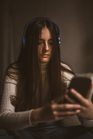 Teen girl excessively sitting at the phone at home. Sad depressed young teenager holding mobile smart phone.の写真素材