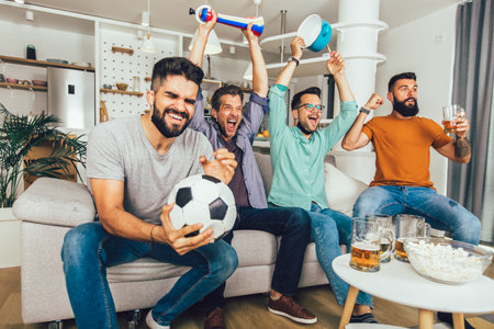 Happy male friends with vuvuzela wearing black face protective mask watching football at homeの写真素材