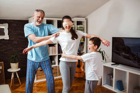 Happy family playing funny hide and seek game at home, blindfolded girl trying to catch her brother.