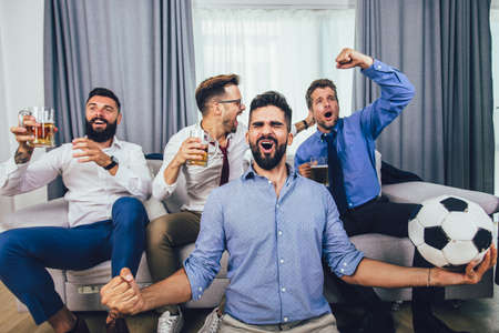 Men watching sport on tv together at home screaming cheerful. Group of friends sitting on the couch and watching a football game.の写真素材