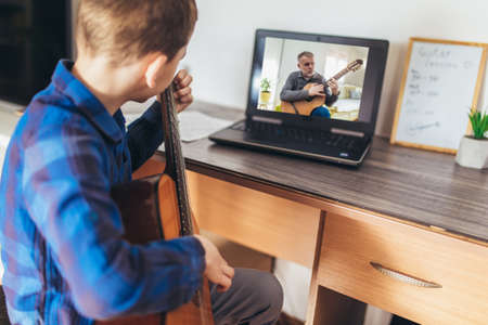 Focused boy playing acoustic guitar and watching online course on laptop while practicing at home. Online training, online classes.の写真素材