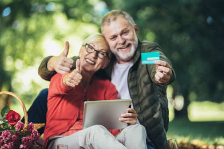 Happy senior couple having a picnic in park using digital tablet and credit card for online shoppingの写真素材