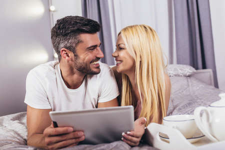 Smiling relaxed young couple using digital tablet in bed at homeの写真素材