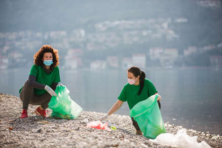Female volunteers clean beach from garbage, plastic, hold green trash bags and care of environment.