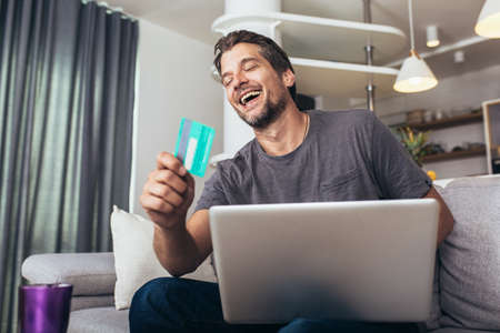 Smiling young handsome man using credit card and laptop at homeの写真素材