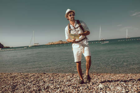 Smiling tourist man standing with map and backpack near the sea.