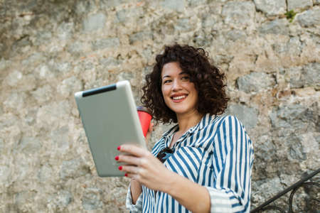 Attractive young woman with curly hair drinking coffee and reading her digital tablet while standing...