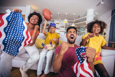 Happy african american family watching rugby match on television in living room at homeの写真素材