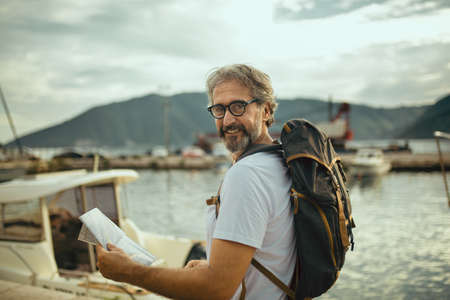 Smiling tourist mature man standing with map and backpack near the sea.の写真素材