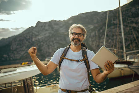 Smiling tourist mature man standing with digital tablet and backpack near the sea.の写真素材