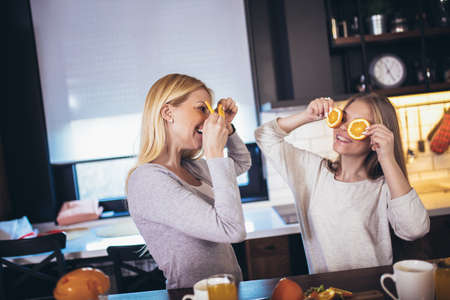 Happy mother and daughter making breakfast at home kitchen and spend time together, having fun with orangeの写真素材