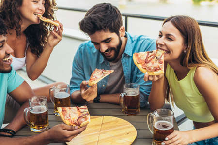Friends enjoying pizza. Group of young cheerful people eating pizza and drinking beer while sitting at the bean bags on the roof of the buildingの写真素材