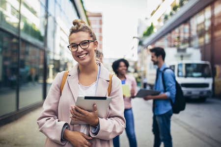 Happy young woman rest and holding books while standing on background of university and friends.の写真素材