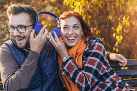 Beautiful young couple sitting on a bench in the autumn park listening to music.の写真素材
