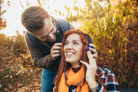 Beautiful young couple sitting on a bench in the autumn park listening to music.の写真素材