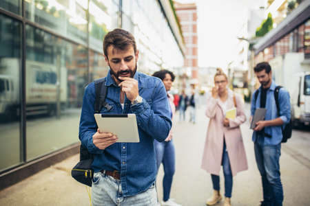 Young man holding digital tablet while standing on background of university and friends.の写真素材