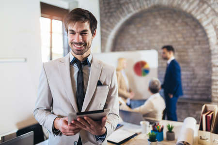 Happy businessman holding digital tablet with team discussing project in the background.