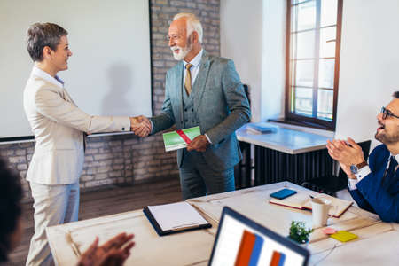 Senior businessman receiving award from businesswoman in front of business professionals, applauding at business seminar in office buildingの写真素材