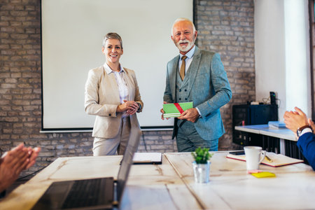 Senior businessman receiving award from businesswoman in front of business professionals, applauding at business seminar in office buildingの写真素材
