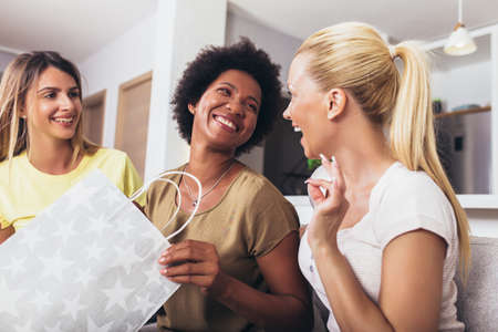 Happy young female friends with wine and shopping bags on sofa at home, having fun after shopping.の写真素材