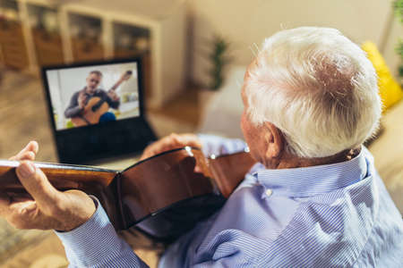 Senior man playing guitar at home using laptop for online lessonsの写真素材